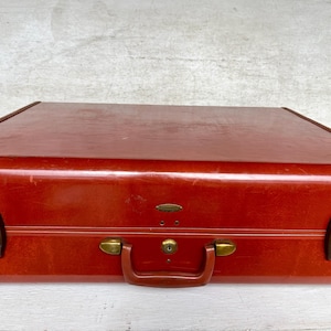 May include: A vintage, reddish-brown suitcase with a glossy finish. The suitcase features a brown handle, brass-colored hardware, and two brown latches on the sides. The suitcase is closed and appears to be in good condition.