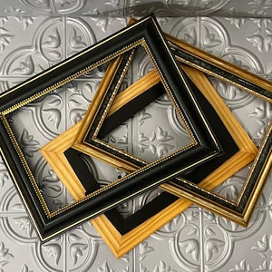 May include: Three empty picture frames in a variety of styles. Two frames have black and gold trim, while the third has a natural wood finish with black accents. The frames are square and arranged on a patterned silver surface.
