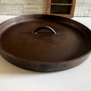 May include: A round, dark brown cast iron lid with a curved handle. The lid has a slightly raised edge and a smooth surface. The handle is a simple, arched design. The lid appears to be well-used and has a rustic, vintage aesthetic.