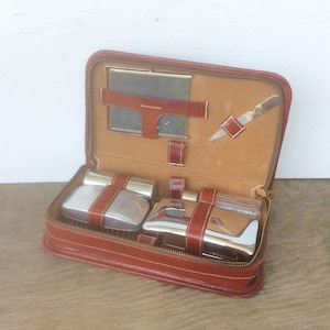 May include: A vintage brown leather travel grooming kit, open to reveal a mirror, small knife, and several silver and glass containers secured with brown leather straps. The kit is zippered and has a tan interior.