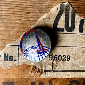 Puede incluir: Un pin de esmalte plateado y azul con un borde rojo y blanco. El pin presenta un diseño estilizado de un edificio con una cúpula y una aguja. El pin está sobre una superficie de madera con un trozo de papel rasgado con el número 201 y el número 96029.