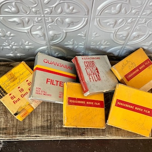 May include: A selection of vintage film boxes, including Kodakchrome and Quantaray brands, in yellow, grey, and red. The boxes are arranged on a weathered wooden surface, suggesting a nostalgic theme for photography.