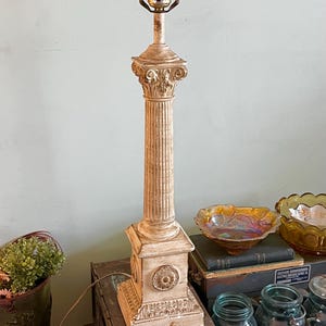 May include: A cream-colored, antique table lamp with a classical column design. The lamp features a fluted column, decorative base, and a bulb socket at the top. The lamp is made of a light-colored material and has an aged appearance.
