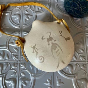 May include: Off-white ceramic pouch with a yellow leather strap. The pouch features gray abstract figures. The pouch is round with a curved top and a leather strap attached at the top. The background is a textured, silver-colored surface.