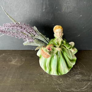 Könnte beinhalten: Keramikfigur einer Frau in einem grünen Kleid, die einen Korb mit rosa Blumen hält. Die Figur ist mit einem Bündel Lavendel und grünem Laub geschmückt. Die Figur hat blondes Haar und trägt ein grünes Kleid.