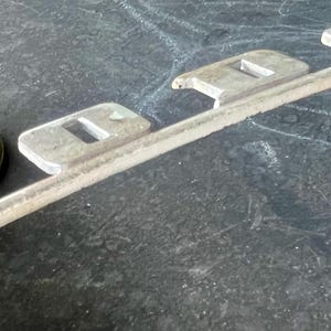 May include: A silver metal car emblem with the word "Dodge" spelled out in raised letters. The emblem is attached to a metal bar.