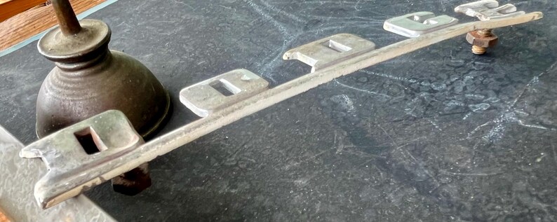 May include: A silver metal car emblem with the word "Dodge" spelled out in raised letters. The emblem is attached to a metal bar.