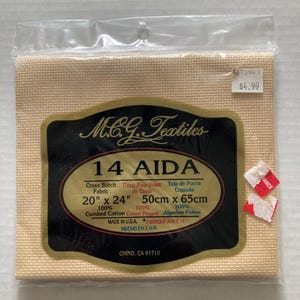 May include: Aida cross stitch fabric, packaged in clear plastic. The label reads "M.C.G. Textiles 14 AIDA" and lists dimensions as 20" x 24". The fabric is a light beige color. Made in the USA.