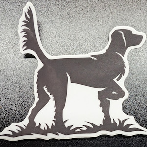 May include: A black and white sticker featuring a silhouette of a dog in a standing pose. The dog has a long tail and is standing on a patch of grass. The sticker has a white border.