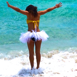 May include: A woman wearing a white tulle tutu skirt and a yellow bikini top stands in the shallow water of a beach.