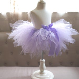 May include: A lavender and white tulle tutu skirt displayed on a mannequin. The skirt has a layered design with a satin bow at the waist. The tutu is on a white stand, set against a neutral-toned background.