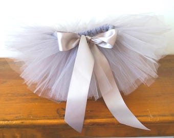 May include: A light gray tulle tutu skirt featuring multiple layers of sheer, gathered fabric. The waistband is elasticized and adorned with a smooth, satin ribbon tied into a centered bow with two long, trailing ends hanging down the front.