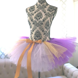 May include: A multi-layered tulle tutu skirt featuring a color palette of deep purple, light lavender, and gold. The skirt is accented with a gold satin ribbon tied in a bow at the center front, displayed on a black and white damask print mannequin.