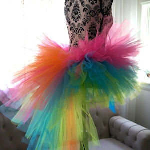 May include: A rainbow tulle tutu skirt with a long train. The skirt is made of multiple layers of tulle in different colours, including pink, orange, yellow, green, blue, and purple. The skirt is on a mannequin.