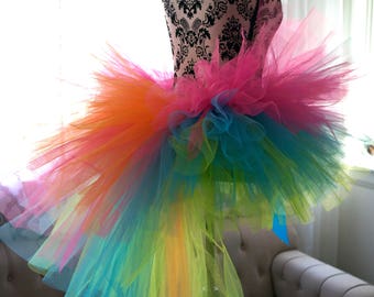 May include: A rainbow tulle tutu skirt with a long train. The skirt is made of multiple layers of tulle in different colors, including pink, orange, yellow, green, blue, and purple. The skirt is on a mannequin.