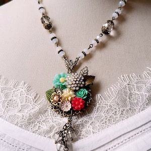 Könnte beinhalten: Eine weiße Halskette mit Kristallperlen und einem floralen Anhänger. Der Anhänger zeigt eine Ansammlung von bunten Blumen, darunter rosa, grüne und weiße, mit einem Kristall in der Mitte. Die Halskette ist mit einem Kristallherz-Anhänger verziert.