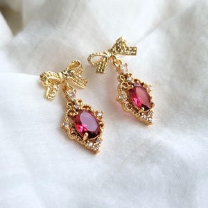 Pode incluir: Um par de brincos de ouro com gotas de pedras preciosas vermelhas e detalhes de laço dourado.