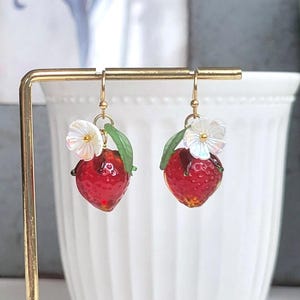 Pode incluir: Um par de brincos de argola de ouro com morangos de vidro vermelho, folhas verdes e flores brancas iridescentes.