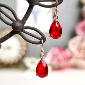 Puede incluir: Un par de pendientes colgantes dorados con grandes piedras preciosas rojas en forma de lágrima. Los pendientes se exhiben en un soporte decorativo de metal oscuro. El fondo está borroso.