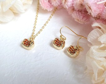 Può includere: Un set di collana e orecchini in oro con un ciondolo e orecchini a forma di cuore. I pezzi a forma di cuore sono ricoperti di piccole pietre preziose arancioni.