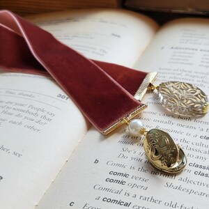 May include: A burgundy velvet bookmark with gold accents and two decorative charms. One charm is a clear glass charm with a gold design, and the other is a gold charm with a heart design. The bookmark is lying on a book with text.