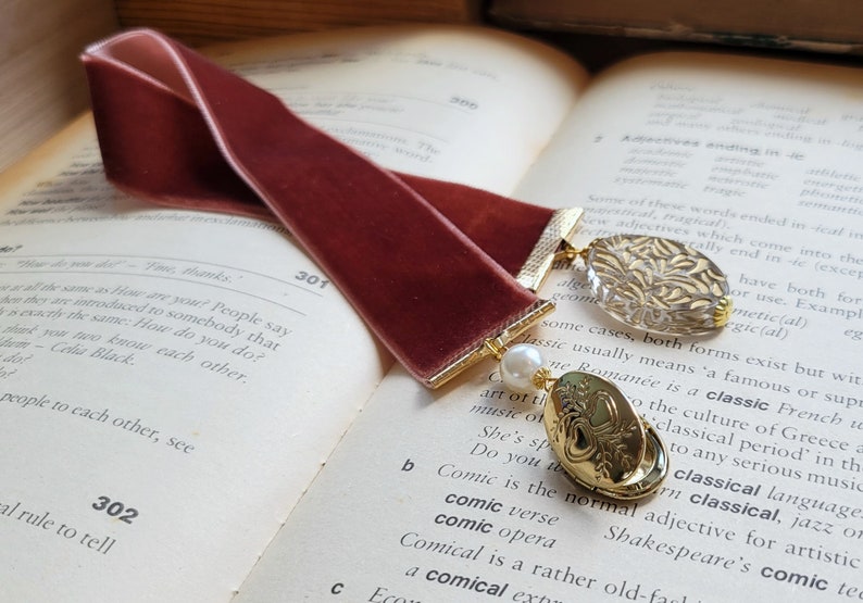 May include: A burgundy velvet bookmark with gold accents and two decorative charms. One charm is a clear glass charm with a gold design, and the other is a gold charm with a heart design. The bookmark is lying on a book with text.