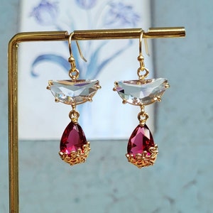 May include: A pair of gold earrings with clear crystal teardrop-shaped pendants and red crystal drops. The earrings have a delicate, feminine design.