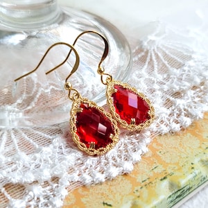 Puede incluir: Un par de pendientes colgantes dorados con grandes piedras preciosas rojas facetadas en forma de lágrima. Las piedras están enmarcadas por un delicado filigrana dorado. Los pendientes cuelgan de ganchos dorados.