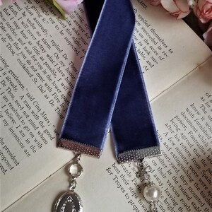 May include: A dark blue velvet ribbon bookmark with silver metal ends and a silver charm with a clear crystal and a silver locket. The bookmark is lying on a book with white pages and pink flowers.