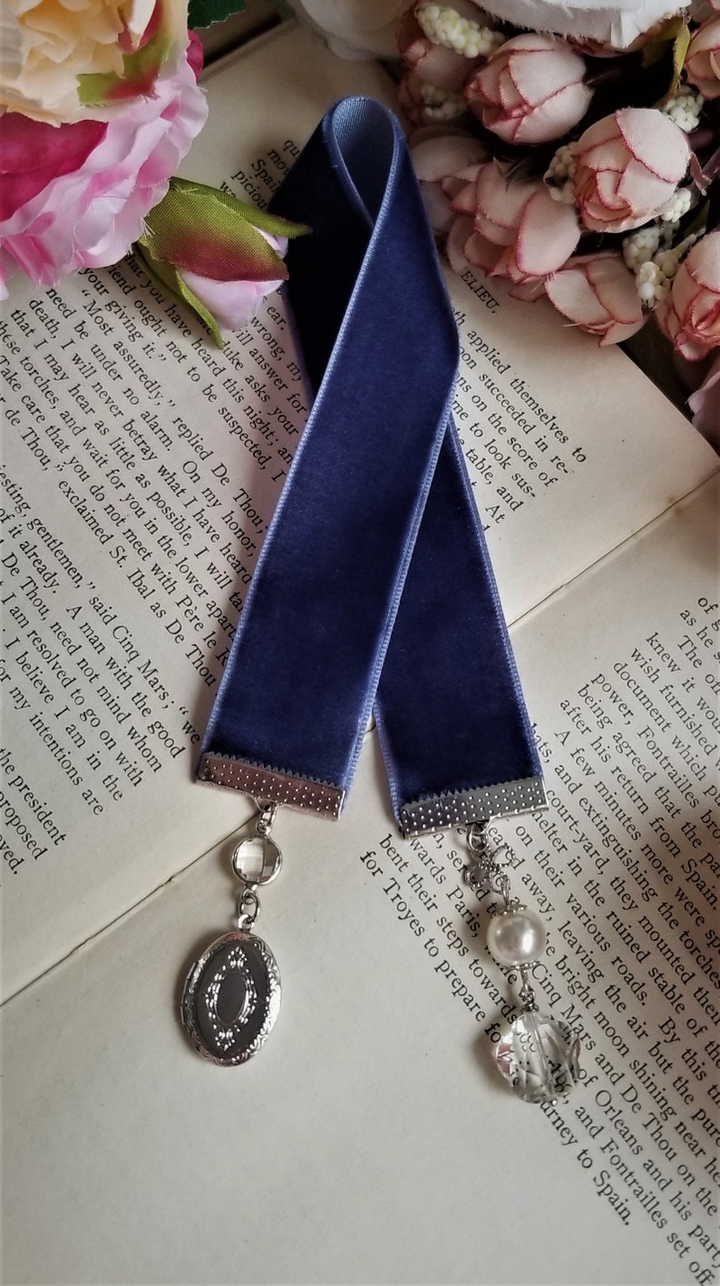 May include: A dark blue velvet ribbon bookmark with silver metal ends and a silver charm with a clear crystal and a silver locket. The bookmark is lying on a book with white pages and pink flowers.