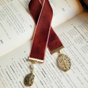 May include: A burgundy velvet bookmark with gold accents and a pearl charm. The bookmark features a gold locket with two hearts and a gold leaf design. The bookmark is resting on a book with text about adjectives ending in -ic.