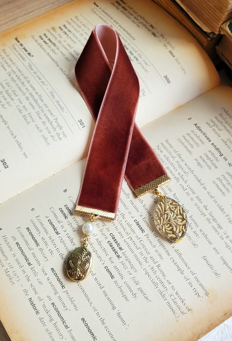 May include: A burgundy velvet bookmark with gold accents and a pearl charm. The bookmark features a gold locket with two hearts and a gold leaf design. The bookmark is resting on a book with text about adjectives ending in -ic.