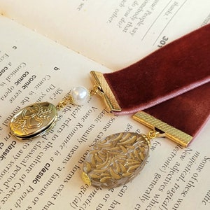 May include: A burgundy velvet bookmark with gold accents and two charms. One charm is a heart-shaped locket with a floral design and the other is a clear charm with a gold floral pattern.