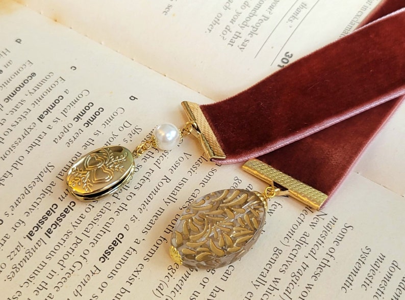 May include: A burgundy velvet bookmark with gold accents and two charms. One charm is a heart-shaped locket with a floral design and the other is a clear charm with a gold floral pattern.