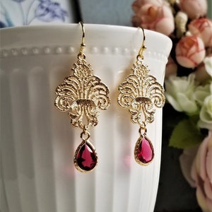 May include: A pair of gold-colored filigree earrings with a teardrop-shaped, ruby-red gemstone. The earrings have a decorative, ornate design and are suspended from a simple hook.