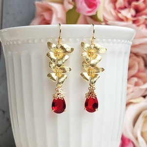 May include: Gold-toned floral drop earrings with a cascading design. Each earring features three flower-shaped elements and a teardrop-shaped, deep red gemstone. The earrings are suspended from a simple hook.