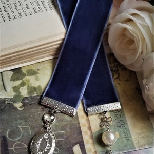May include: A blue velvet bookmark with silver metal accents. The bookmark has a locket charm with a floral design and a crystal charm.