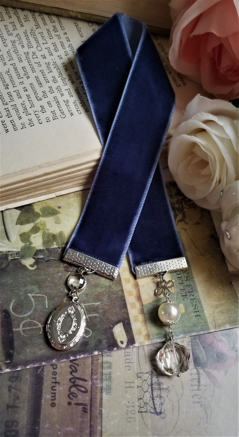 May include: A blue velvet bookmark with silver metal accents. The bookmark has a locket charm with a floral design and a crystal charm.