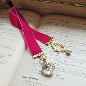 Puede incluir: Un marcapáginas de terciopelo rosa con un dije dorado en forma de corona y una perla. El marcapáginas está sobre un libro abierto con el texto "split infinitive" visible.