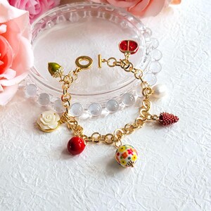 Puede incluir: Un brazalete de cadena dorada con dijes, incluyendo una mariquita roja, una rosa blanca, una baya roja, una bellota verde, una cuenta roja y una cuenta blanca con flores de colores.