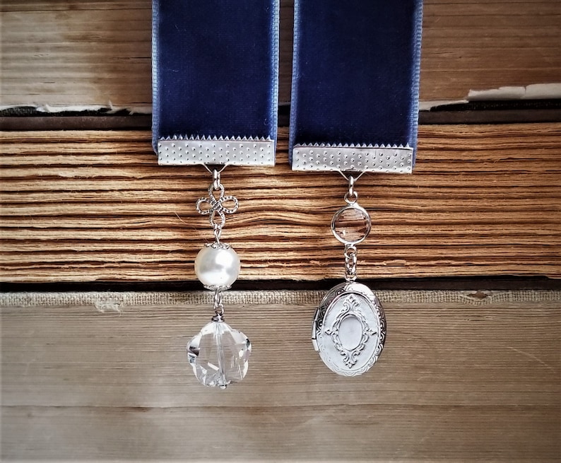 May include: Two blue velvet ribbon bookmarks with silver charms. One charm is a pearl and crystal pendant, the other is a silver locket with a crystal.