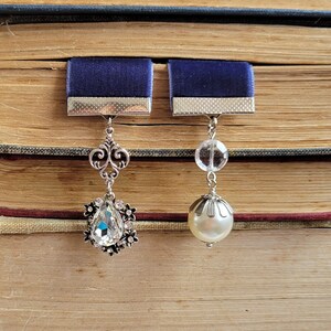 May include: Two silver bookmark charms with blue velvet ribbon accents. One charm features a clear crystal teardrop and the other a white pearl.