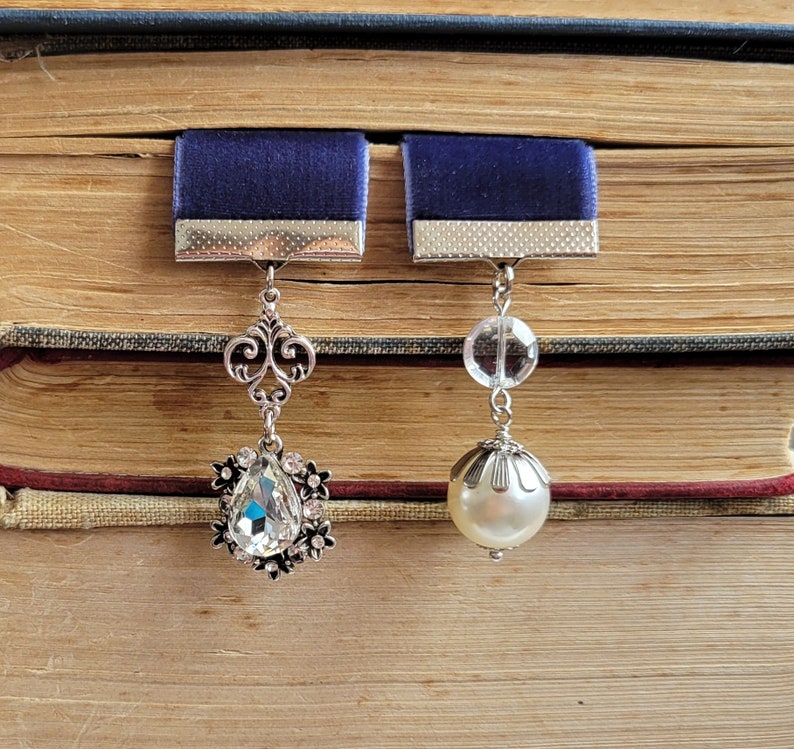 May include: Two silver bookmark charms with blue velvet ribbon accents. One charm features a clear crystal teardrop and the other a white pearl.