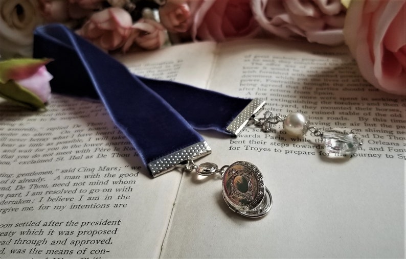 May include: A dark blue velvet bookmark with a silver chain and charm. The charm features a pearl and a crystal. The bookmark is lying on a book with text.