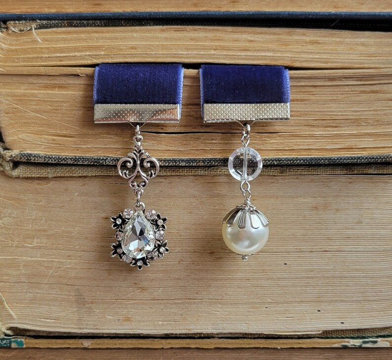 May include: Two silver bookmark charms with blue velvet ribbon accents. One charm features a teardrop-shaped crystal and the other a pearl.