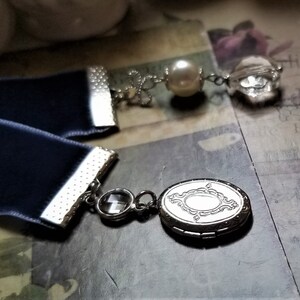 May include: A navy blue velvet ribbon bookmark with silver accents and a silver locket charm. The locket has a decorative design and a clear crystal charm hangs from the ribbon.