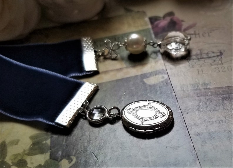 May include: A navy blue velvet ribbon bookmark with silver accents and a silver locket charm. The locket has a decorative design and a clear crystal charm hangs from the ribbon.