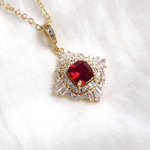 May include: A gold necklace with a square, ruby-red gemstone pendant. The pendant is encircled by a halo of small, clear crystals and baguette-cut stones. The chain is fine, and the jewellery is set against a white backdrop.