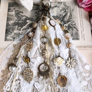 Könnte beinhalten: Ein handgefertigter Quastenanhänger mit weißer Spitze, goldfarbenen Perlen und verschiedenen Anhängern. Das Design umfasst ovale und florale Anhänger, Perlen und eine Blütenperle an der Spitze. Die Quaste ist mit einem Metallverschluss versehen.