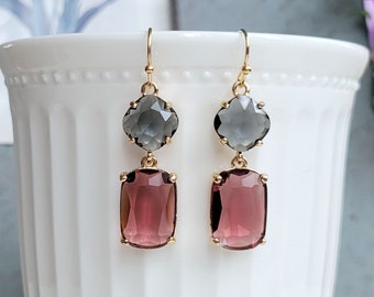 May include: A pair of gold-toned drop earrings. Each earring features a gray, four-leaf clover-shaped stone above a rectangular, reddish-purple stone. The earrings are displayed against a white background.